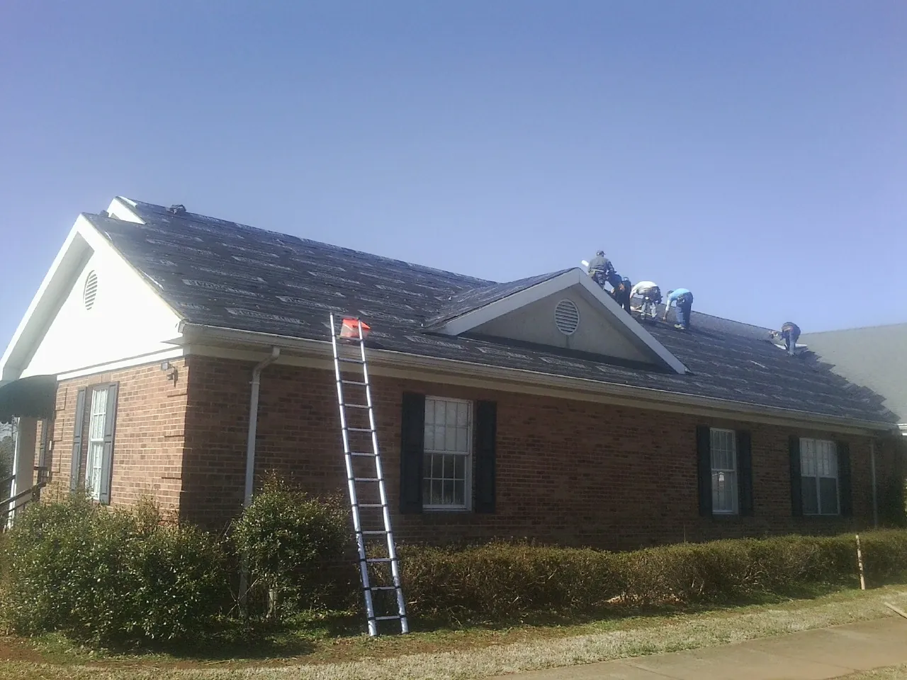 Crew tearing shingles off a brick ranch roof with a ladder set — Emergency Roof Repair work by our crew in Ridgeland