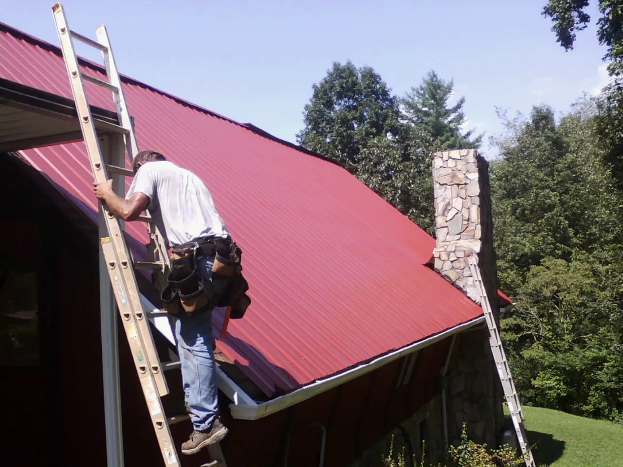 Roofer on a ladder beside a red metal roof and stone chimney — Soffit & Fascia Repair by Roofing Brothers in Ridgeland